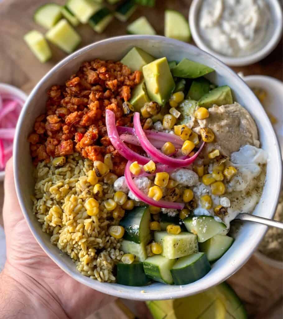 holding a Mediterranean ground chicken bowl with pickled onion, avocado, roasted corn, tzatziki, and rice