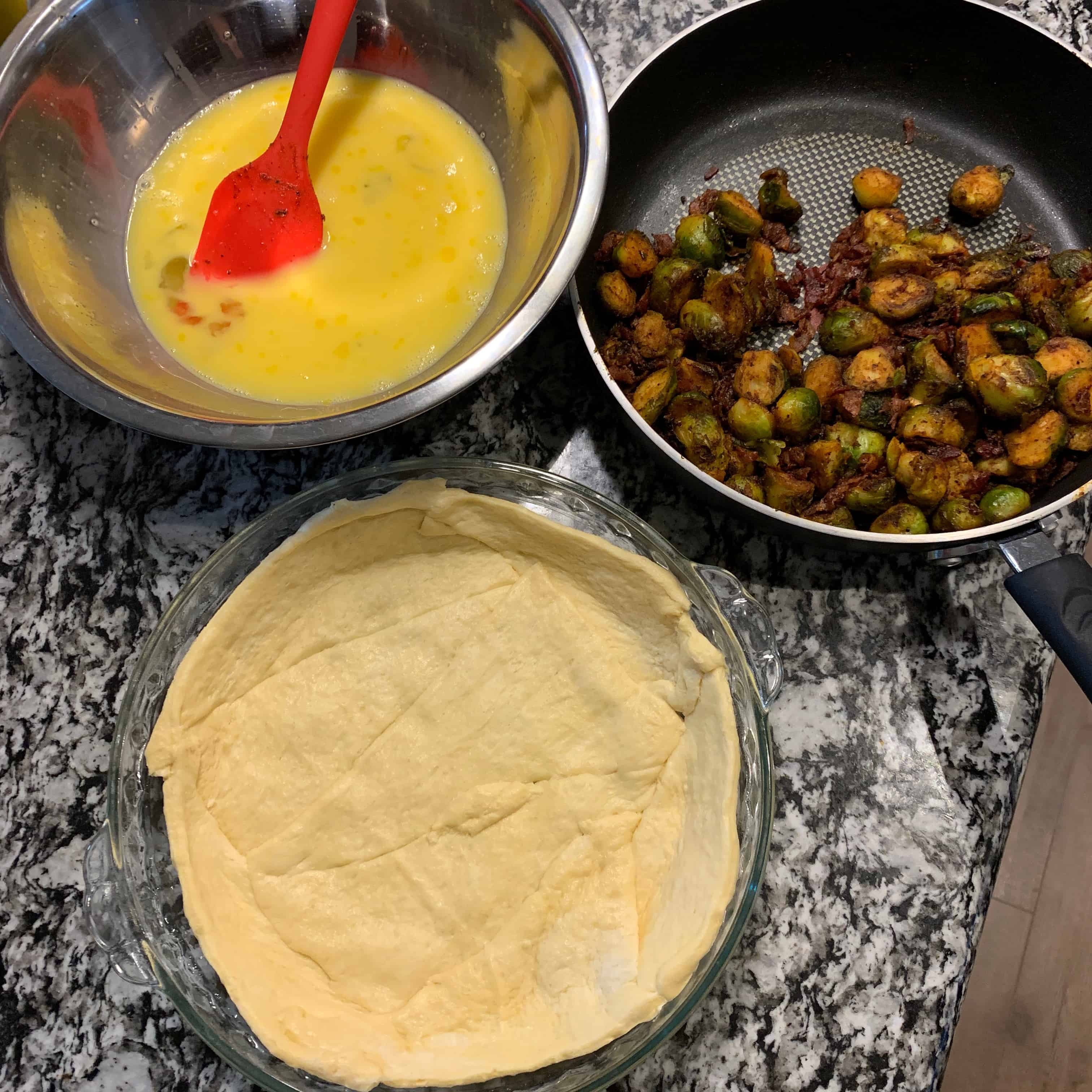 crescent rolls rolled flat in a pie dish, smoked brussels sprouts in a pan with bacon, and whisked eggs in a bowl before assembling the breakfast pie