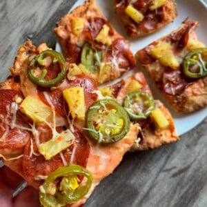 holding a piece of low calorie pepperoni pizza over a plate with 4 more slices