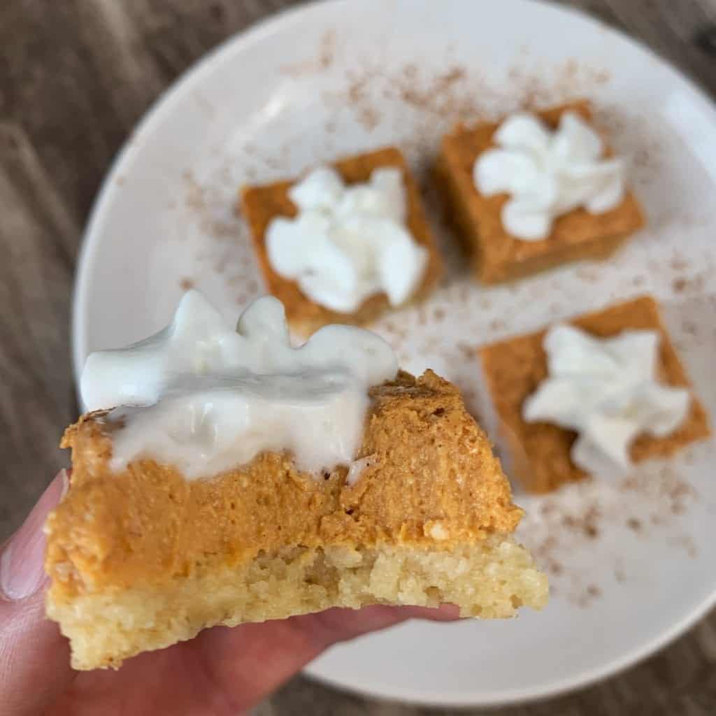 pumpkin cheesecake bar with a bite taken out of it and whipped cream on top with 3 more pumpkin cheesecake bars in the background on a plate