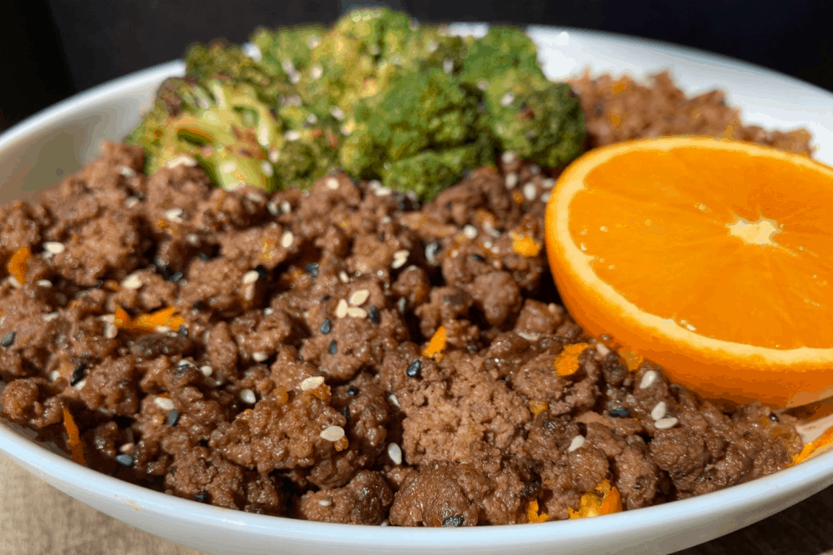 crispy orange beef in a white bowl with broccoli, fried cauliflower rice, and half an orange
