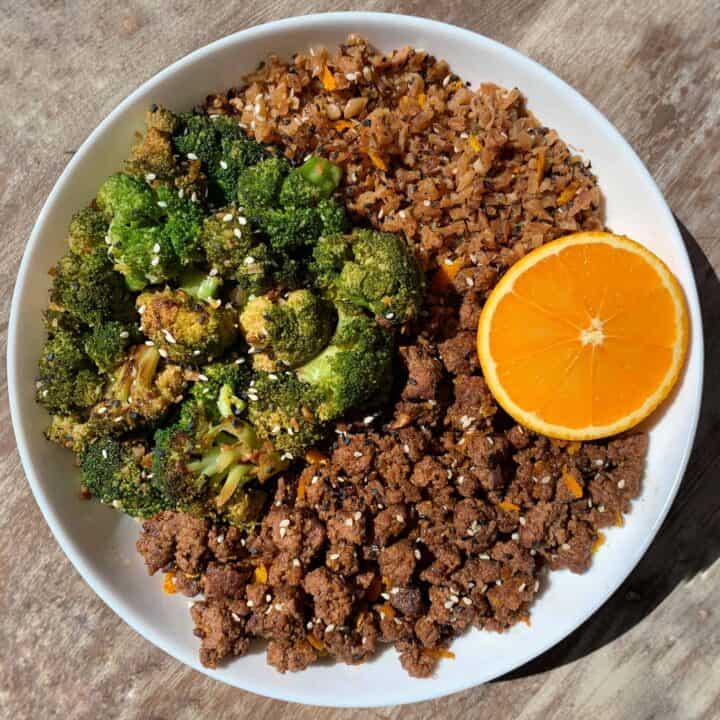 crispy orange beef in a white bowl with broccoli, fried cauliflower rice, and half an orange
