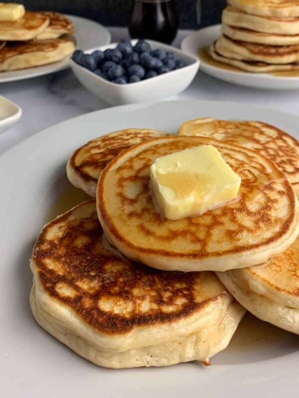 stack of protein powder pancakes with butter and syrup in front of more pancakes, syrup, and a bowl of blueberries