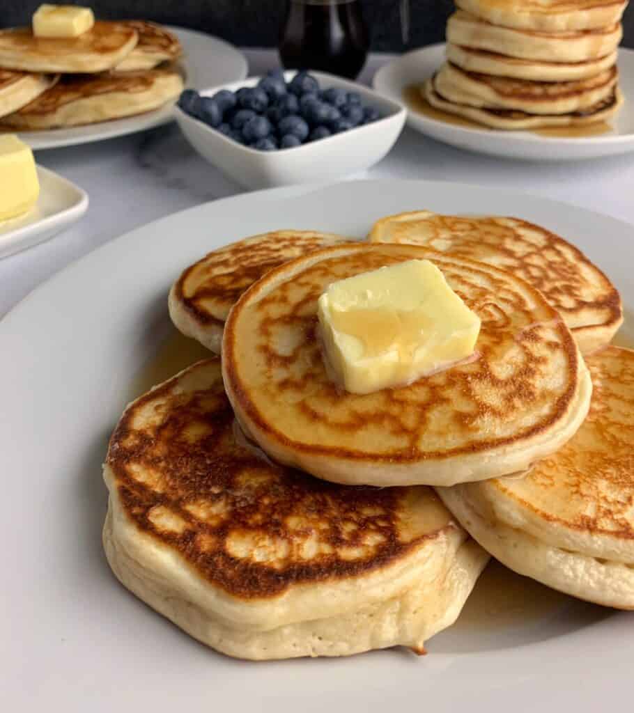stack of protein powder pancakes with butter and syrup in front of more pancakes, syrup, and a bowl of blueberries