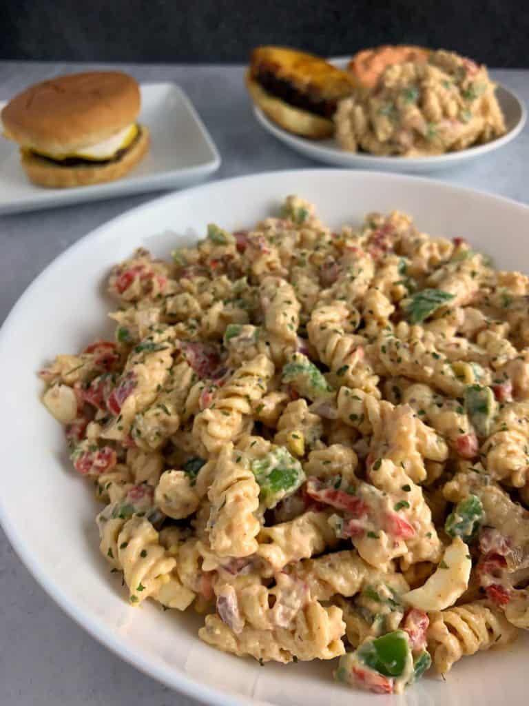 healthy pasta salad in a white bowl with burgers in the background