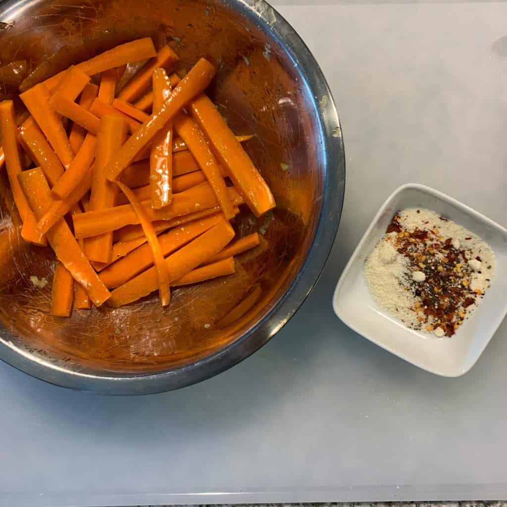 sliced carrot fries in a mixing bowl coated in garlic infused olive oil with a bowl of grated parmesan, red pepper, and black pepper on the side