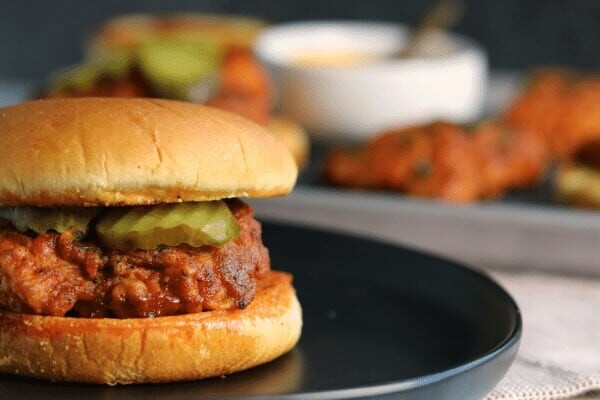 chicken sandwich on a black plate with a tray of pan fried chicken, buns, and sauce in the background