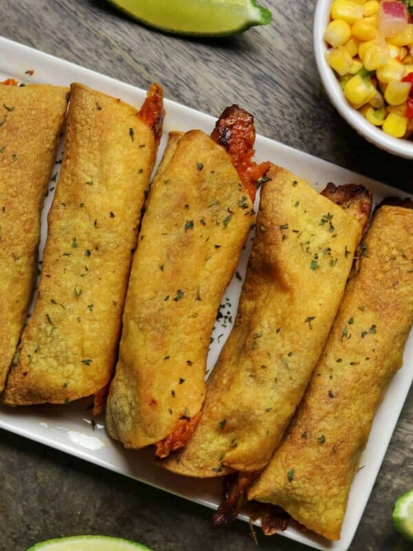 chicken taquitos on a plate with corn salsa, guacamole, and lime wedges