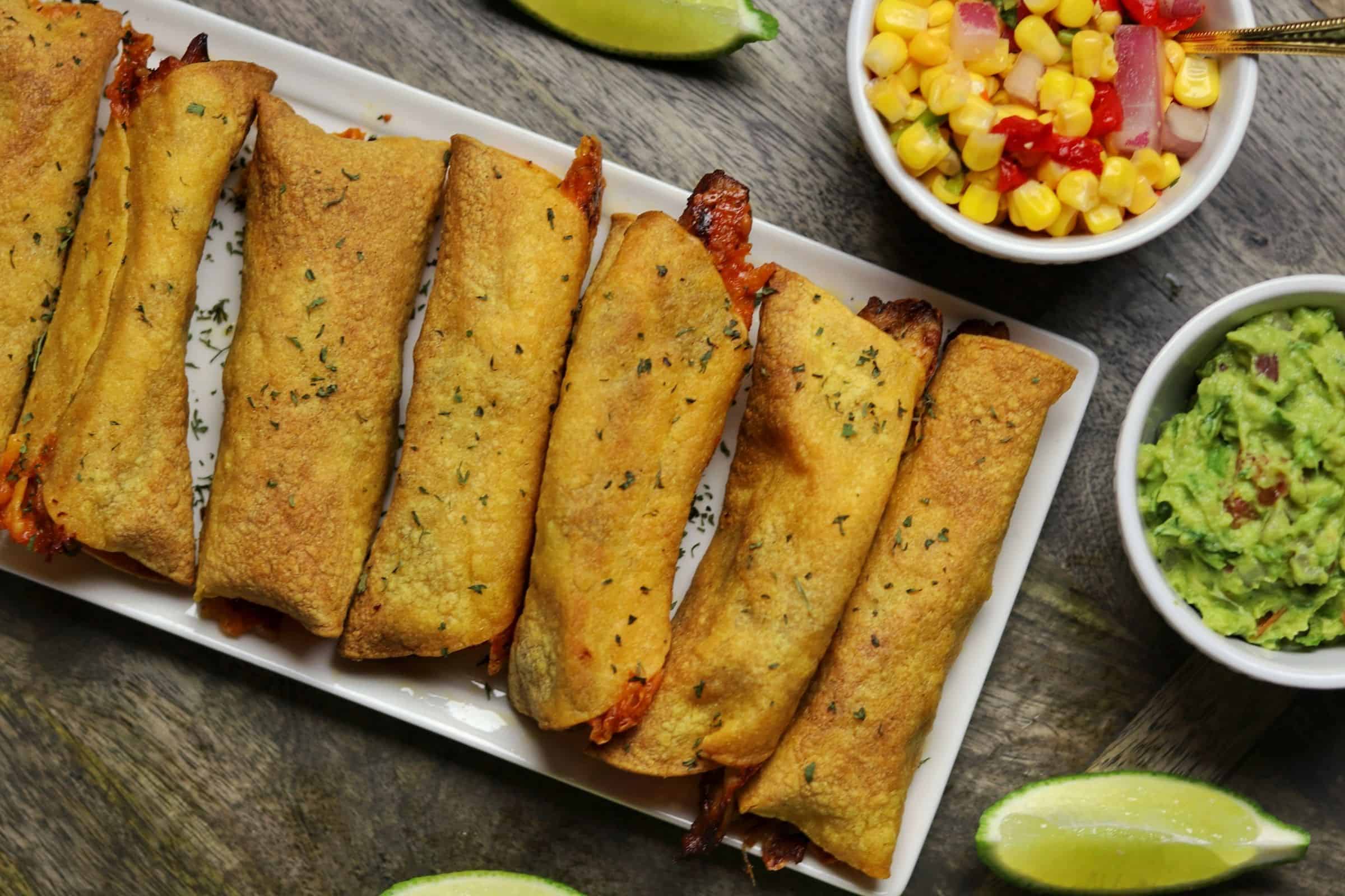 chicken taquitos on a plate with corn salsa, guacamole, and lime wedges