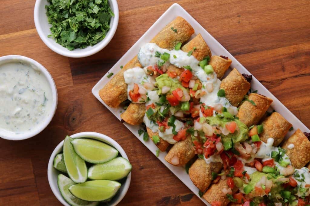 a white plate with baked taquitos topped with guacamole, fat free Greek yogurt, cilantro, and pico de gallo