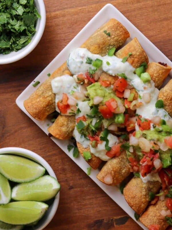 a white plate with baked taquitos topped with guacamole, fat free Greek yogurt, cilantro, and pico de gallo