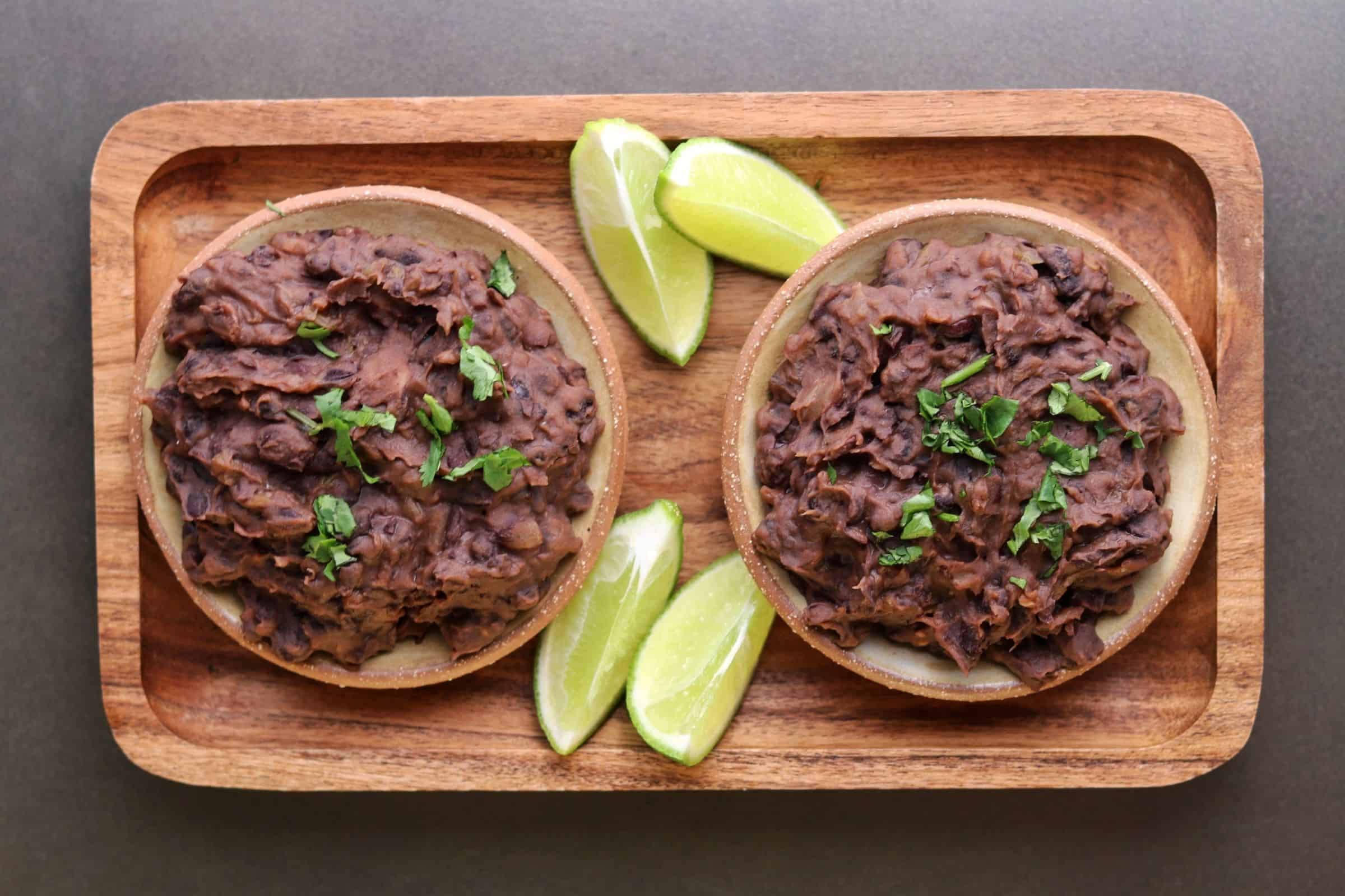 two bowls of refried black beans with lime wedges and cilantro