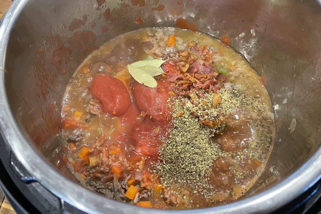 cooked bacon, peeled tomatoes, dried oregano, bay leaves, and red cooking wine added to the ground beef