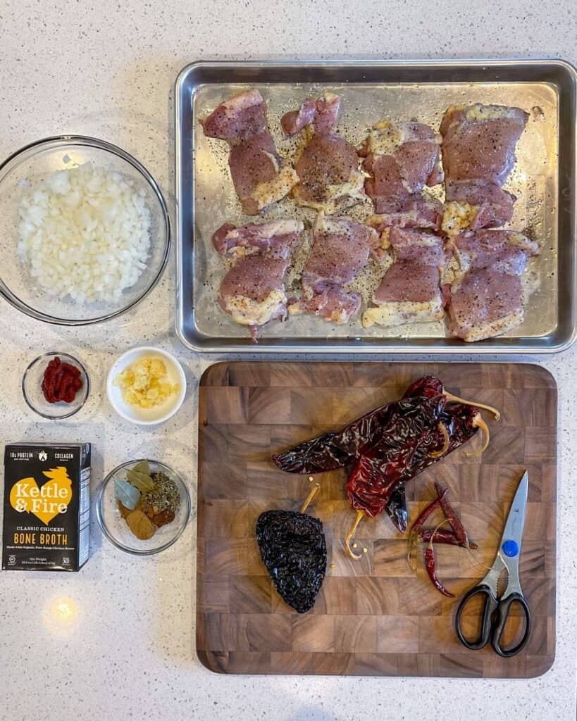 dried guajillo, ancho, and arbol chiles on a cutting board with kitchen scissors next to a sheet pan with seasoned boneless skinless chicken thighs, diced white onion, grated garlic, tomato paste, chicken bone broth, and a bowl of spices and bay leaves