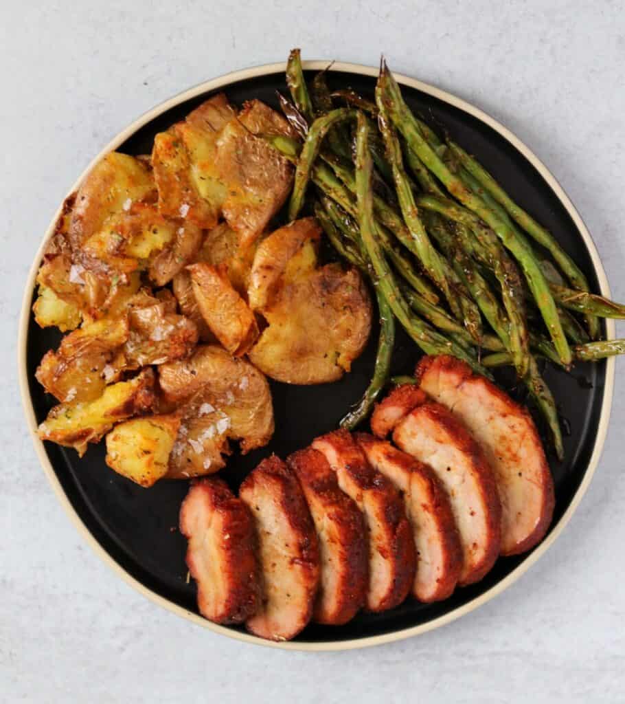 sliced pork tenderloin on a black plate with green beans and smashed potatoes