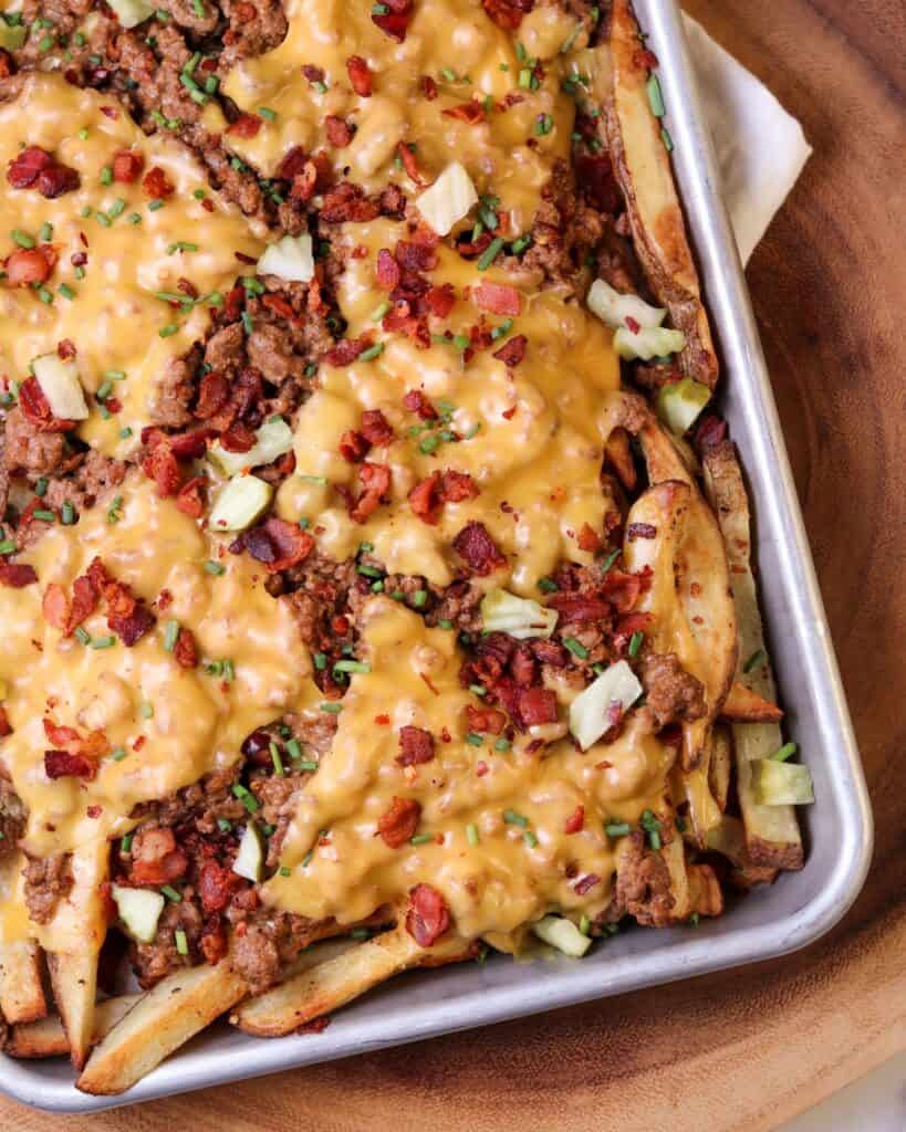 quarter sheet pan of cheeseburger fries topping with bacon, diced pickles, and chives