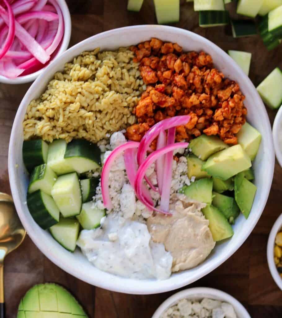 honey harissa ground chicken in a bowl with rice, goat cheese, cucumbers, avocado, hummus, tzatziki, and pickled onion