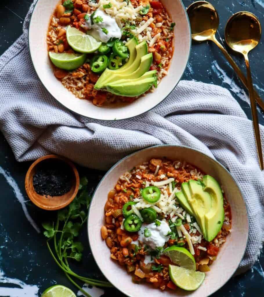 two bowls of chili topped with avocado slices, cheese, jalapeño, and lime wedges
