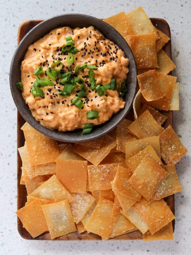 baked wonton chips on a wooden plate with a bowl of chicken dip topped with black sesame seeds and scallions