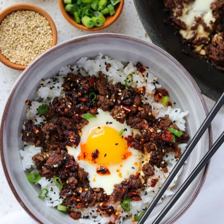 ground beef and eggs in a bowl with rice, green onion, sesame seeds, and chili crisp