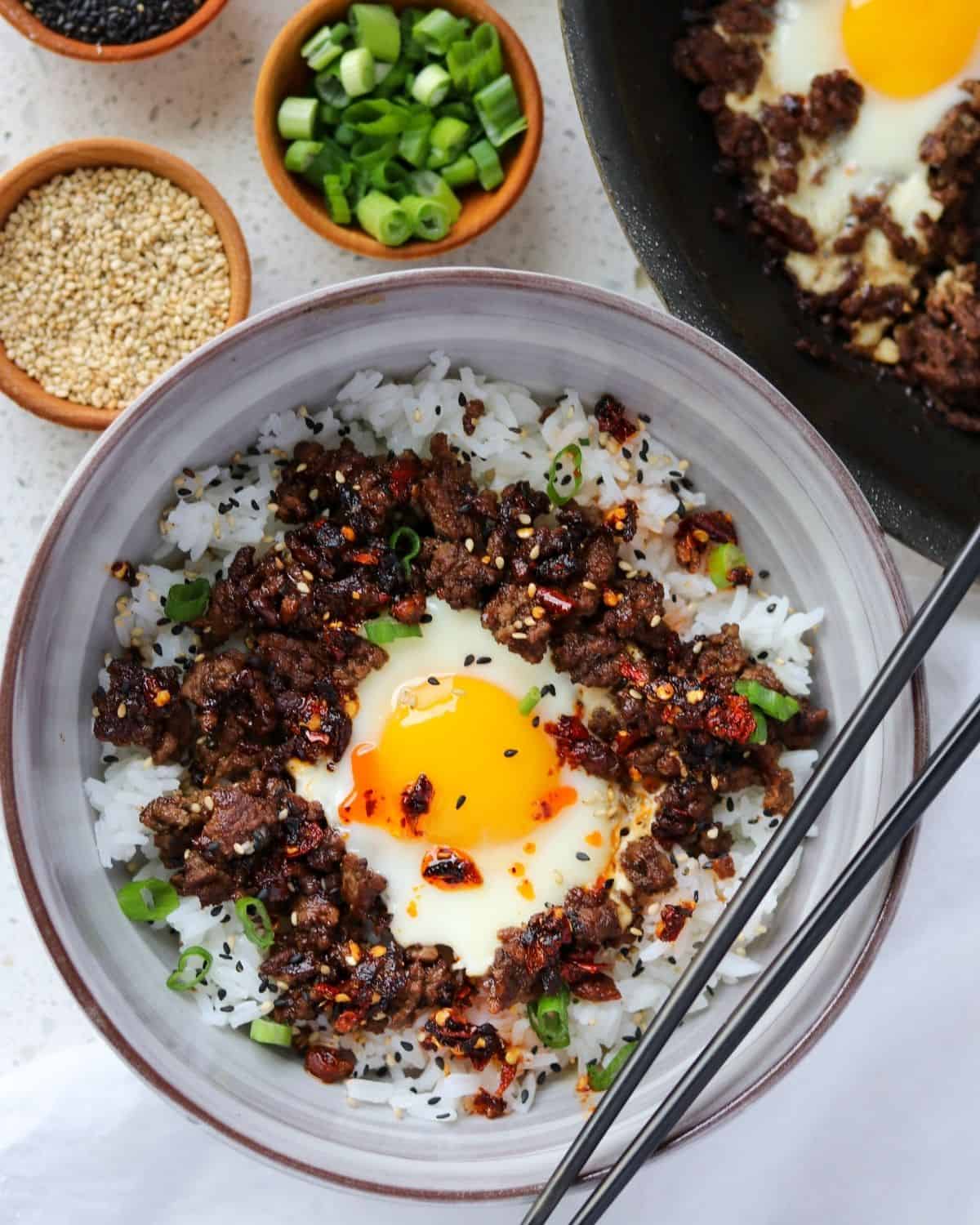 ground beef and eggs in a bowl with rice, green onion, sesame seeds, and chili crisp