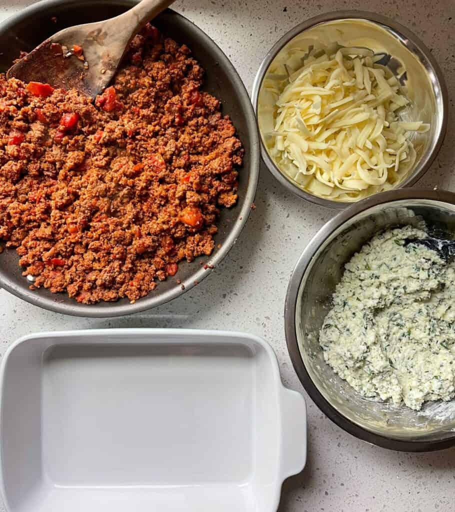 meat sauce in a skillet, mozzarella in a bowl, and cheese sauce in a bowl next to a baking dish