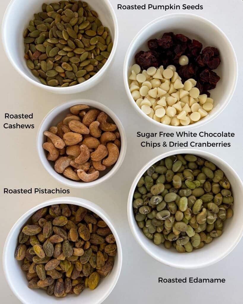 roasted edamame, pistachios, cashews, and pumpkin seeds in bowls beside a bowl of white chocolate chips and dried cranberries