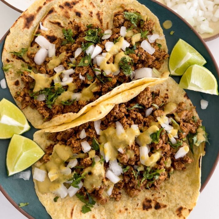 two ground pork tacos on a plate topped with diced onion, cilantro, salsa, and lime wedges