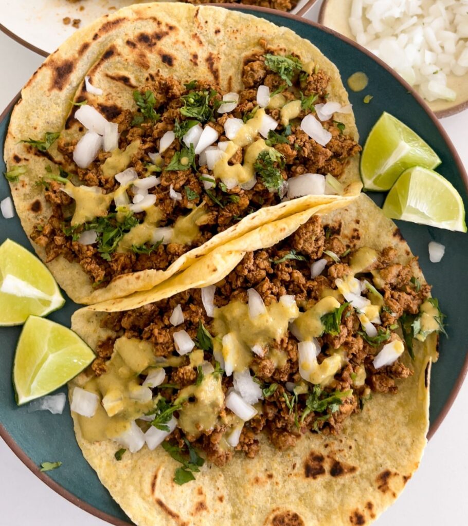two ground pork tacos on a plate topped with diced onion, cilantro, salsa, and lime wedges