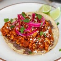ground chicken tinga tostada with refried black beans, pickled onions, cilantro, queso fresco, and lime wedges