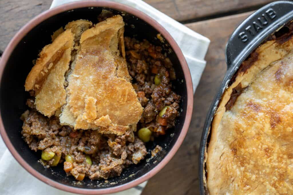 serving of ground beef pot pie in a bowl beside the whole pie in a cast iron skillet
