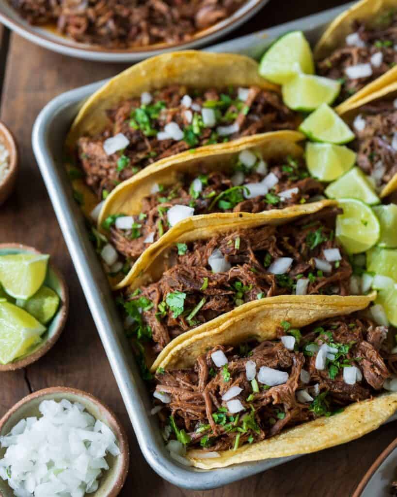 pulled beef tacos in a sheet pan with lime wedges and garnishes of diced onion, cilantro, and cotija cheese