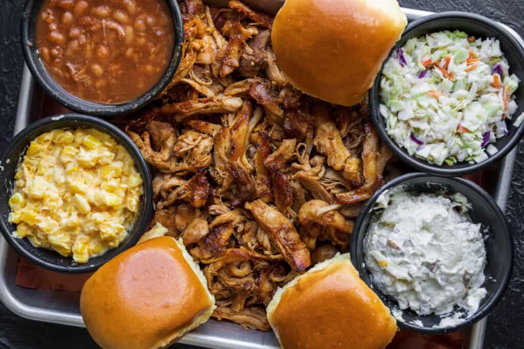 bbq shredded chicken on a sheet pan with rolls, baked beans, potato salad, cole slaw, and corn