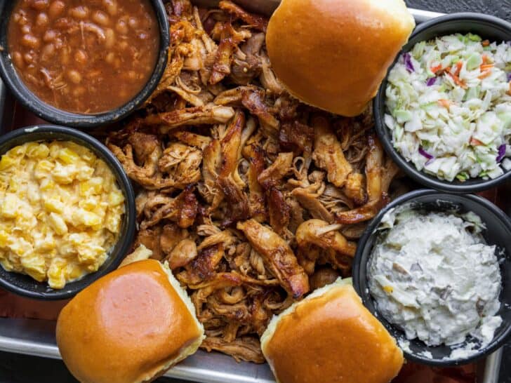 bbq shredded chicken on a sheet pan with rolls, baked beans, potato salad, cole slaw, and corn