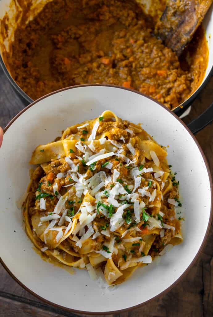 holding a bowl of pumpkin bolognese with pappardelle above a pot of bolognese 