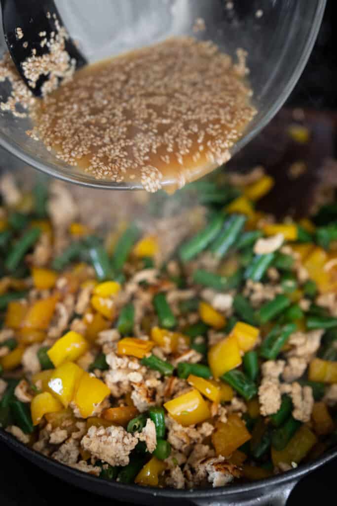 pouring a honey sesame sauce on cooked ground chicken, bell pepper, and green beans