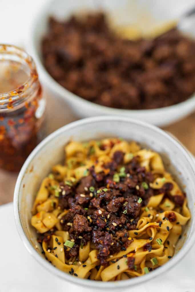 bowl of noodles with firecracker beef in front of a jar of chili oil and a bowl of spicy ground beef