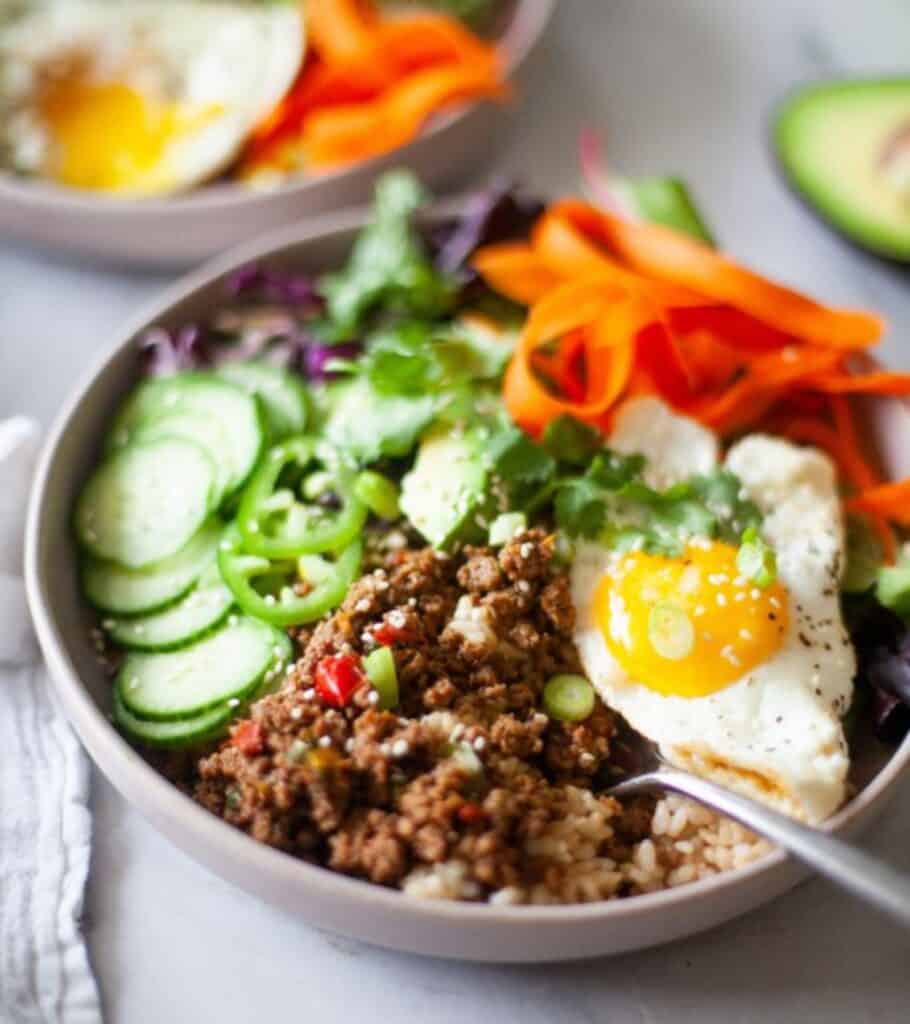 bowl with Korean beef, sliced cucumber, carrots, fried egg, cabbage, and rice