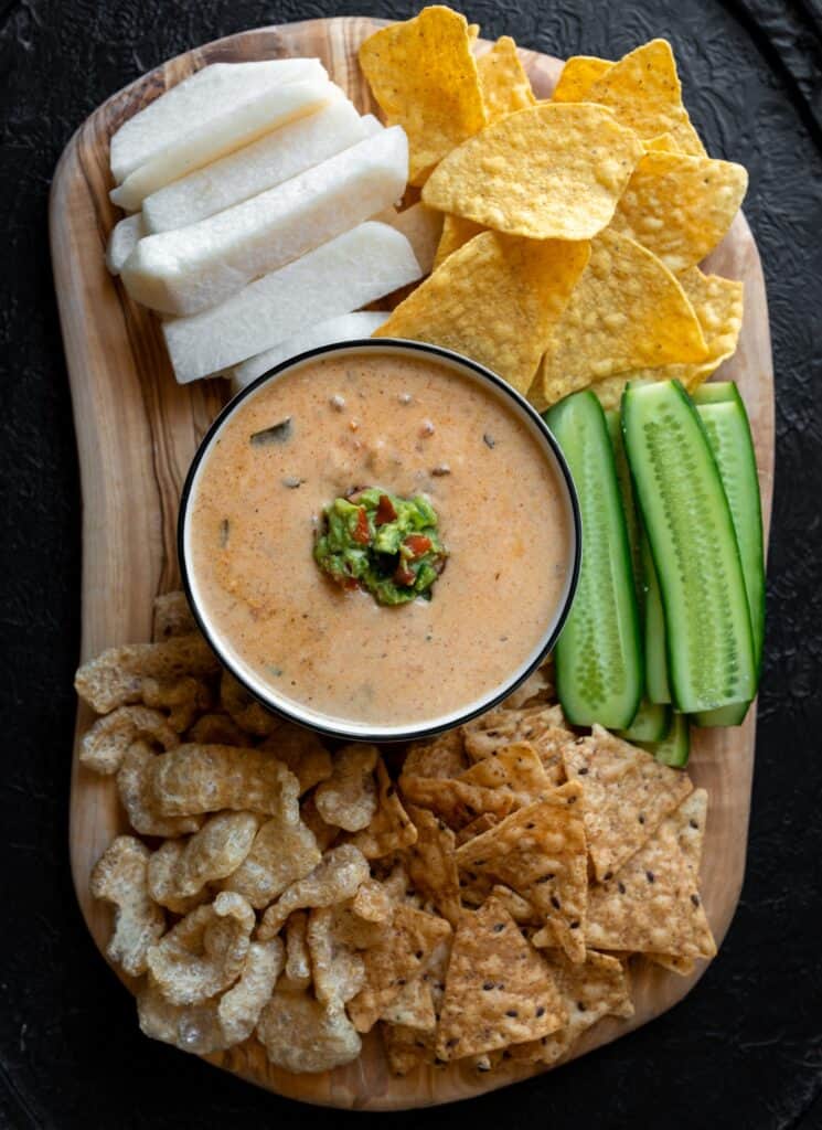 protein queso on a board with sliced cucumber, pork rinds, tortilla chips, and jicama chips