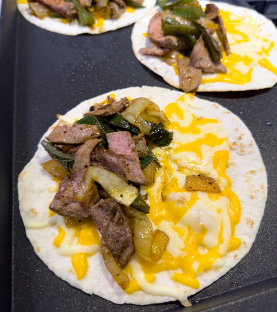 tortillas on a griddle topped with shredded cheese, steak, peppers, and onion