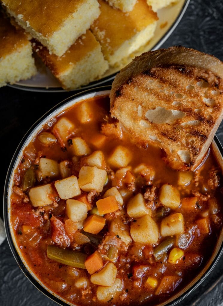 bowl of hamburger soup with a sliced of grilled sourdough bread in it and a plate with cornbread behind it