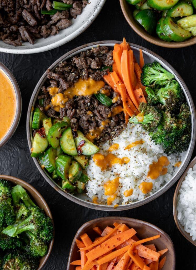 ground beef bulgogi bowl with kimchi sauce, sticky rice, sesame seeds, roasted broccoli, cucumber salad, and pickled carrots