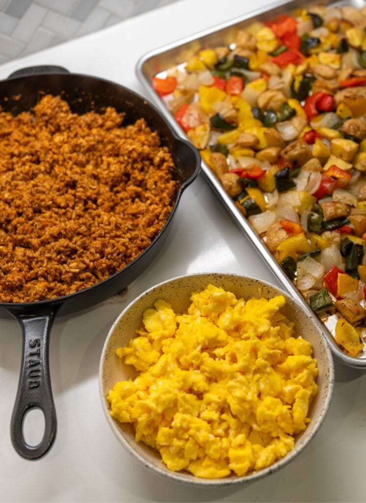 cooked ground chicken in a skillet next to a bowl of scrambled eggs and a sheet pan with roasted potatoes, peppers, and onions
