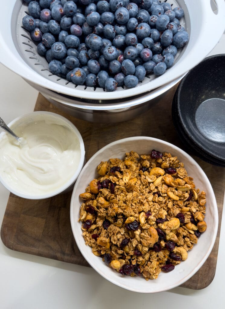 fresh blueberries in a colander, vanilla Greek yogurt, and a bowl of homemade granola 