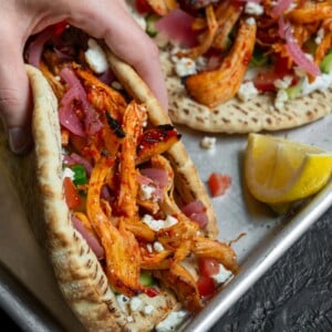 holding a gyro with honey harissa chicken, feta, tzatziki, cucumber, tomato, and pickled onion