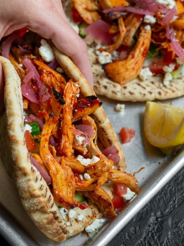 holding a gyro with honey harissa chicken, feta, tzatziki, cucumber, tomato, and pickled onion
