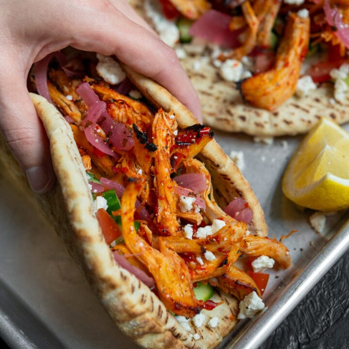 holding a honey harissa chicken gyro with pickled onion, tzatziki, feta, cucumber, and tomato