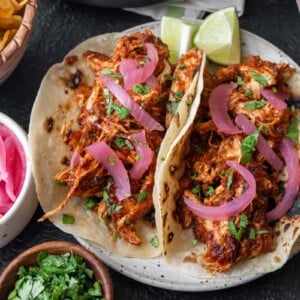 two rotisserie chicken tacos on a plate with lime wedges, pickled onions, and cilantro