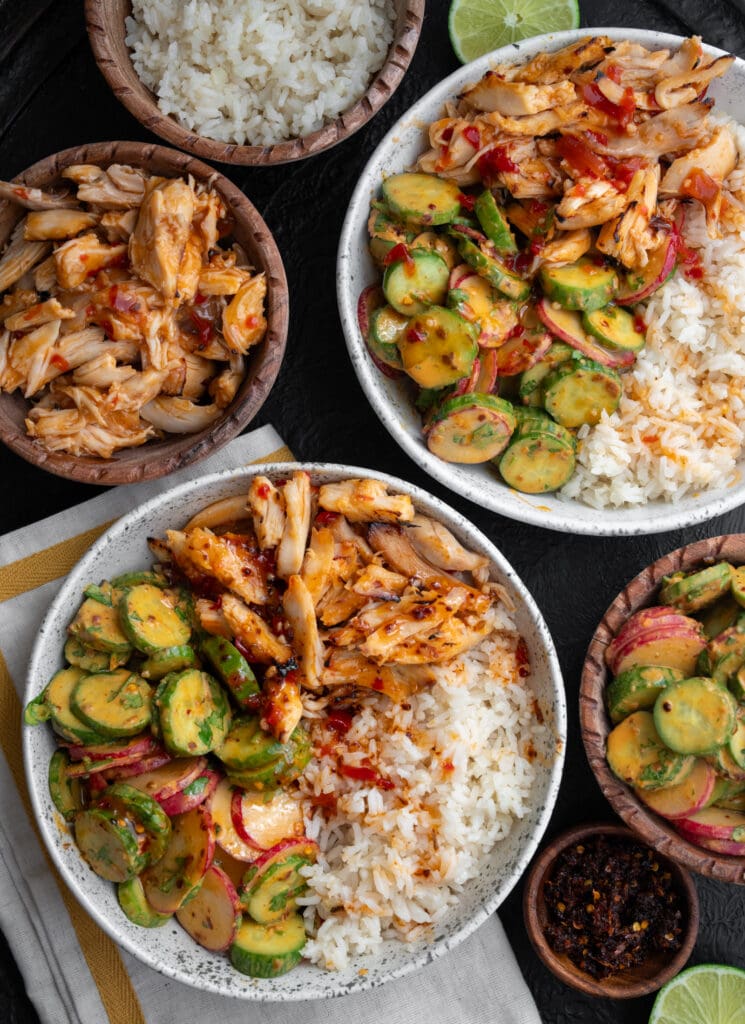 two rotisserie chicken bowls with bowls of rice, cucumber crunch, chili crisp, sweet chili chicken, and lime halves