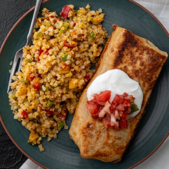 cheesy rotisserie chicken burrito on a plate with Mexican cauliflower rice garnished with Greek yogurt and salsa roja